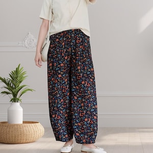 May include: A woman wearing a white v-neck top and black pants with a floral print. She is standing in a room with white walls and a plant in a white vase.