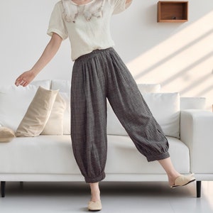 May include: A woman wearing a white blouse with a crocheted detail and black and white checkered pants. She is standing in a room with a white couch and a potted plant.