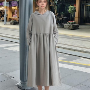 Pode incluir: Um vestido cinza com capuz, cintura franzida e mangas compridas. O vestido está sendo usado por uma pessoa em pé em uma calçada em frente a um prédio.