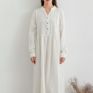 May include: A white long-sleeved button-down dress with a v-neckline. The dress has a gathered skirt and falls to the mid-calf. The model is wearing white sneakers.