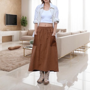 May include: A woman wearing a brown corduroy skirt with two large front pockets, a white crop top, and a striped button-down shirt. She is also wearing beige boots. The skirt is a midi length.