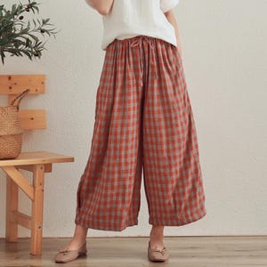 May include: A woman wearing a white short-sleeved top and wide-leg, plaid pants in shades of red, brown, and gray. The pants have a drawstring waist. The woman is standing in front of a light-colored wall.