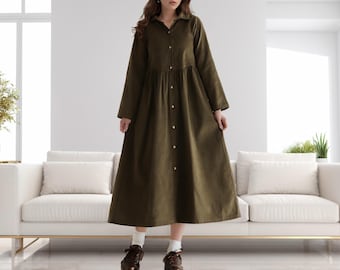 May include: An olive green long-sleeve dress with a button-down front and a collared neckline. The dress has a gathered waist and a flowing skirt. The model is wearing brown shoes and white socks. The dress is made of a soft, corduroy-like fabric.
