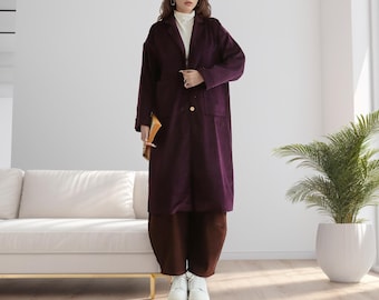 May include: A long, purple corduroy coat with a single button closure and a pocket on the left side. The coat is worn by a person with brown hair and is standing in a room with a white couch and a potted plant.