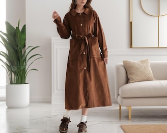 May include: A brown corduroy trench coat with a button closure, a belt, and two patch pockets. The coat is worn by a person with brown hair and brown eyes. The person is standing in a white room with a white couch and a round mirror in the background.
