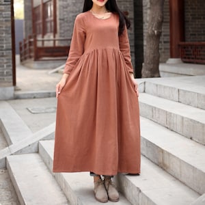May include: A long, rust-colored linen dress with a round neckline and long sleeves. The dress has a gathered waist and falls to the ankles. The model is wearing the dress with brown ankle boots.