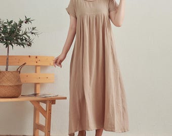 Könnte beinhalten: Eine Frau trägt ein hellbraunes, locker sitzendes, knielanges Kleid mit kurzen Ärmeln. Das Kleid hat einen runden Ausschnitt und einen gerafften Rock.