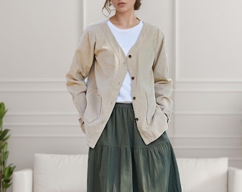 Könnte beinhalten: Eine Frau trägt einen Strohhut, eine beige Strickjacke, ein weißes T-Shirt und einen langen, dunkelgrünen Rock. Das Outfit ist lässig und stilvoll, passend für warmes Wetter. Die Strickjacke hat Taschen und Knöpfe. Der Rock ist bodenlang.