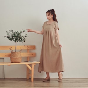 Puede incluir: Una mujer lleva un vestido largo de color marr&oacute;n claro, holgado, de manga corta y cuello redondo. Est&aacute; de pie frente a un banco de madera con una planta en maceta.