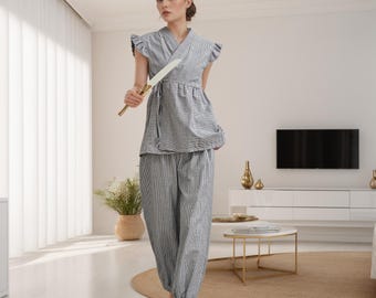Puede incluir: Una mujer lleva una blusa gris y blanca a rayas y pantalones a juego, sostiene un cuchillo blanco frente a ella. Está de pie en una habitación con una pared blanca y un televisor en la pared.
