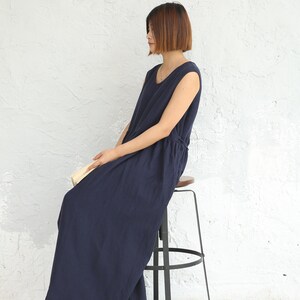 May include: A woman wearing a navy blue sleeveless maxi dress sits on a metal stool with a wooden seat. The dress has a drawstring waist and a relaxed fit.