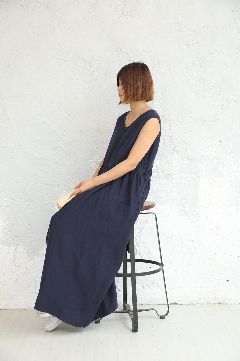 May include: A woman wearing a navy blue sleeveless maxi dress sits on a metal stool with a wooden seat. The dress has a drawstring waist and a relaxed fit.