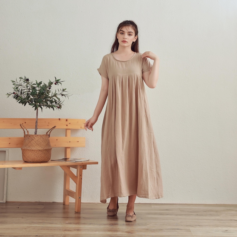 Puede incluir: Una mujer lleva un vestido de color beige claro, holgado, de largo hasta la rodilla, con mangas cortas. El vestido tiene un escote redondo y una falda fruncida.