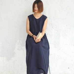 May include: A woman wearing a navy blue sleeveless maxi dress sits on a chair with a book in her lap. She has short brown hair and is looking down.