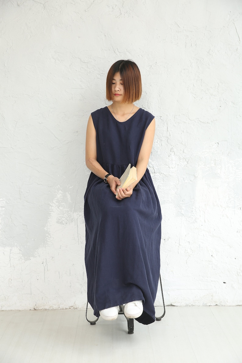 May include: A woman wearing a navy blue sleeveless maxi dress sits on a chair with a book in her lap. She has short brown hair and is looking down.