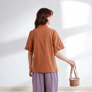 Pode incluir: Uma mulher vestindo uma camisa marrom de manga curta e cal&ccedil;as roxas est&aacute; segurando uma pequena cesta de vime com uma al&ccedil;a branca.