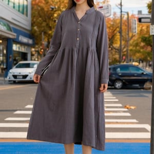 May include: A woman wearing a long-sleeved, gray, button-down dress with a gathered skirt. She is standing on a crosswalk in front of a building.