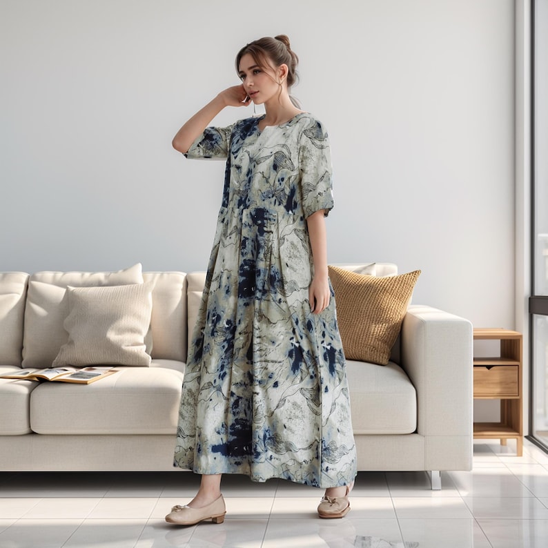 May include: A woman wearing a long, flowing dress with a blue and white abstract print. The dress has a round neckline and short sleeves. The woman is standing in front of a white sofa with a tan cushion.