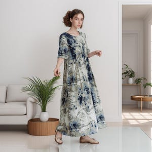 May include: A woman wearing a long, flowing dress with a blue and white abstract print. The dress has a round neckline and three-quarter sleeves. She is standing on a white floor with a plant and a sofa in the background.