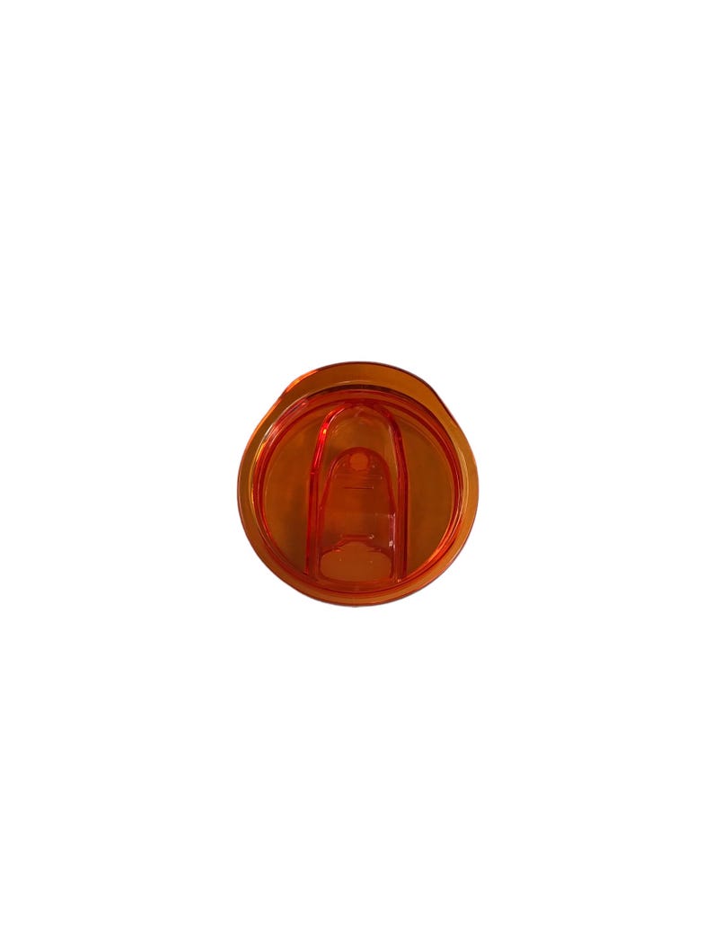 Op de afbeelding: Een oranje plastic tumblerdeksel met een schuifsluiting. Het deksel is cirkelvormig met een verzonken midden en een klein lipje om de schuif te openen en te sluiten. Het plastic is doorschijnend.