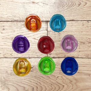 Op de afbeelding: Een bovenaanzicht toont negen kleurrijke, transparante tumblerdeksels in verschillende tinten: oranje, teal, paars, rood, roze, geel, groen en blauw. De deksels zijn gerangschikt op een lichtbruin houten oppervlak.