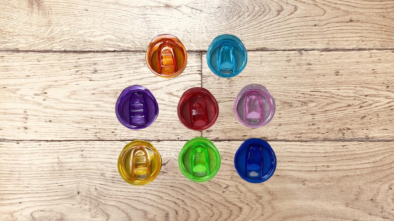 Op de afbeelding: Een bovenaanzicht toont negen kleurrijke, transparante tumblerdeksels in verschillende tinten: oranje, teal, paars, rood, roze, geel, groen en blauw. De deksels zijn gerangschikt op een lichtbruin houten oppervlak.