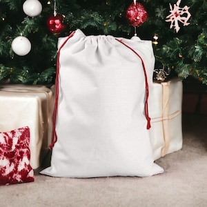 May include: A white drawstring gift bag with red cord, standing in front of a decorated Christmas tree. Presents wrapped in brown paper and a red and white patterned pillow are visible. Ornaments in red, white, and silver adorn the tree.