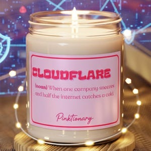 Puede incluir: Una vela encendida en un tarro de cristal con una etiqueta rosa que dice "CLOUDFLARE" y "Pinktionary". La vela está sobre un pequeño posavasos de madera, con luces de cadena y un fondo borroso.