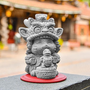 May include: A gray stone figurine of a lion wearing a crown and holding a small turtle. The figurine is sitting on a red base.