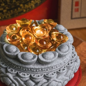 Könnte beinhalten: Eine graue Steinschale mit aufwendigen Schnitzereien, gefüllt mit einem Haufen glänzender Goldbarren. Die Schale ist ein traditionelles chinesisches Symbol für Reichtum und Wohlstand.