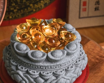 May include: A grey stone bowl with intricate carvings, filled with a pile of shiny gold ingots. The bowl is a traditional Chinese symbol of wealth and prosperity.
