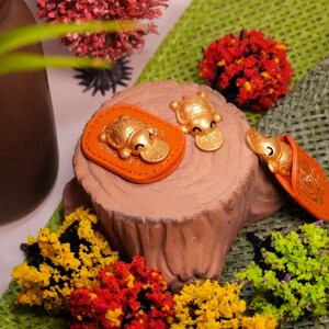 Könnte beinhalten: Goldfarbene Schildkrötenfiguren mit Münzen, präsentiert auf orangefarbenen Lederaufnähern und einem Baumstumpf. Die Szene ist mit bunten Blumen und Grün dekoriert und schafft eine lebendige und glückverheißende Darstellung.