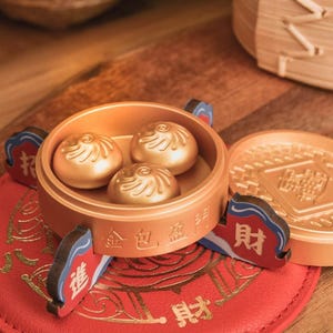 May include: A golden-colored dim sum set with three buns inside a round container. The container sits on a red mat with gold Chinese characters. The set includes a lid and decorative supports with Chinese characters.