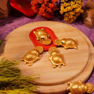 Può includere: Una collezione di statuette di tartarughe dorate, alcune con monete, disposte su una superficie di legno. È presente anche un pezzo rettangolare rosso con un disegno di tartaruga. La scena è decorata con verde e accenti colorati.