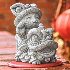 May include: A gray stone sculpture of a cat riding a lion. The cat is wearing a hat and has a playful expression. The lion is detailed with intricate carvings.