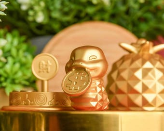 May include: A gold-coloured figurine of a turtle holding a gold coin with a dollar sign on it. The turtle is sitting on a gold-coloured base with a gold-coloured pineapple next to it.