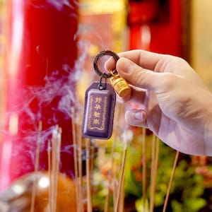 May include: A purple leather keychain with gold text and a small gold-colored bell charm. The keychain is held in a hand, with incense and a red background visible.