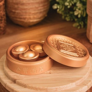 May include: A gold-colored, round container with three gold ingots inside, and a lid with a detailed design. The container sits on a wooden surface, with text in Chinese characters. The overall theme is related to wealth and prosperity.