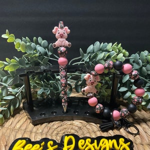 Op de afbeelding: Een zwarte en roze pen met een bloemenpatroon en een roze varkensbedel bovenop. Een zwarte en roze armband met een bloemenpatroon en een roze varkensbedel. Een zwart bord met gele tekst dat "Bee's Designs" zegt.