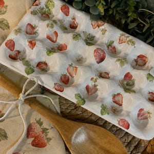 May include: A white ceramic egg carton featuring a repeating pattern of red strawberries and green leaves. The carton has twelve molded indentations for eggs. A wooden spoon and a beige fabric cloth with a matching strawberry print are visible.