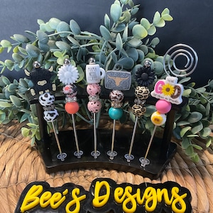 以下が含まれることがあります： 6本のカラフルなビーズで飾られた装飾的な棒のセットで、それぞれに異なるチャームが付いています。チャームには、牛、花、コーヒーカップ（「ママはコーヒーが必要」の文字入り）、牛の頭蓋骨、ひまわり、そして「神と共にすべてが可能である」の文字入りのひまわりがあります。棒は、「Bee's Designs」という黄色の文字が書かれた黒いスタンドに飾られています。
