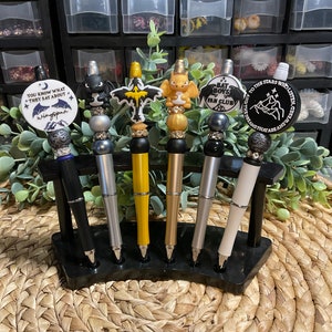 May include: Five decorative pens with different colored tops and charms. The charms include a dragon, a bat, a winged creature, a cartoon bat boy, and a mountain scene. The pens are displayed in a black pen holder.