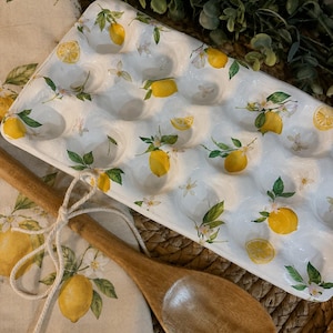 Op de afbeelding: Een witte keramische eierhouder met een herhalend patroon van gele citroenen, groene bladeren en witte bloesems. Een houten lepel is met een wit touwtje vastgebonden aan een bijpassende linnen doek met een citroen- en bloesemprint.