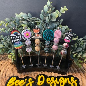 Könnte beinhalten: Ein schwarzer Ständer mit sechs silbernen Metallstäben, an denen bunte Perlen-Schlüsselanhänger befestigt sind. Die Schlüsselanhänger zeigen verschiedene Designs, darunter einen Regenbogen, ein Smiley, einen Fuchs, einen türkisfarbenen Kreis mit dem Text "Zero Fucks Club", einen rosa-weißen Regenbogen und einen Totenkopf mit dem Text "Drop Dead". Der Ständer steht auf einer gewebten Oberfläche mit dem Text "Bee's Designs" in Gelb.