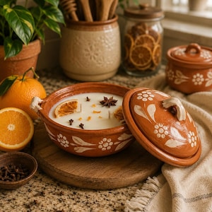 May include: A terracotta clay pot candle with a lid, featuring white hand-painted floral patterns. The white wax is topped with dried orange slices, star anise, and whole cloves. The pot has two small handles and sits on a round wooden board.