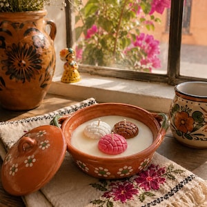 May include: A terracotta bowl with a lid, featuring hand-painted white flower accents, filled with white wax and three small, round, textured candle inserts in white, brown, and pink, each with a central wick, resting on a floral embroidered cloth.