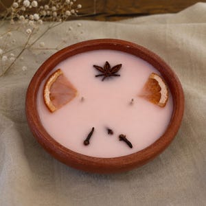 May include: A round terracotta bowl filled with pale pink wax, featuring two wicks. The surface is decorated with two dried orange slices, one star anise, and three whole cloves embedded into the wax, creating a textured and fragrant appearance.