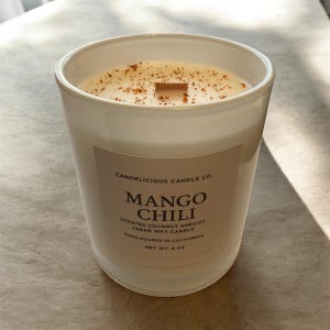 May include: A white glass candle with the text "Mango Chili" on the label. The candle has a creamy wax with a wooden wick and is sprinkled with small brown particles. The net weight is 8 oz.