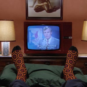 Könnte beinhalten: Ein Paar Socken mit einem sich wiederholenden geometrischen Muster aus orangefarbenen, roten und schwarzen Sechsecken. Die Socken werden an Füßen getragen, die vor einem alten Fernsehgerät mit einer Nachrichtensendung und dem Text 10 GLENN RINKER WPLG Miami positioniert sind.