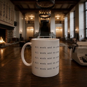 May include: White ceramic mug with the text "All work and no play" repeated in black. The mug has a curved handle and is on a wooden surface. A vintage typewriter is visible in the background.